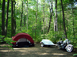 Site de campement rustique en forêt avec une tente, une motocyclette et une remorque.