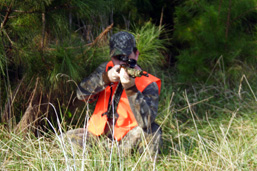 Un homme en habit de camouflage avec une veste orangée est accroupi dans l'herbe et pointe son arme à feu dans la direction de l'objectif.